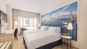 Habitación de hotel con una cama grande, un escritorio, una silla, una mesita con una planta y un cuadro de un paisaje costero en la pared.