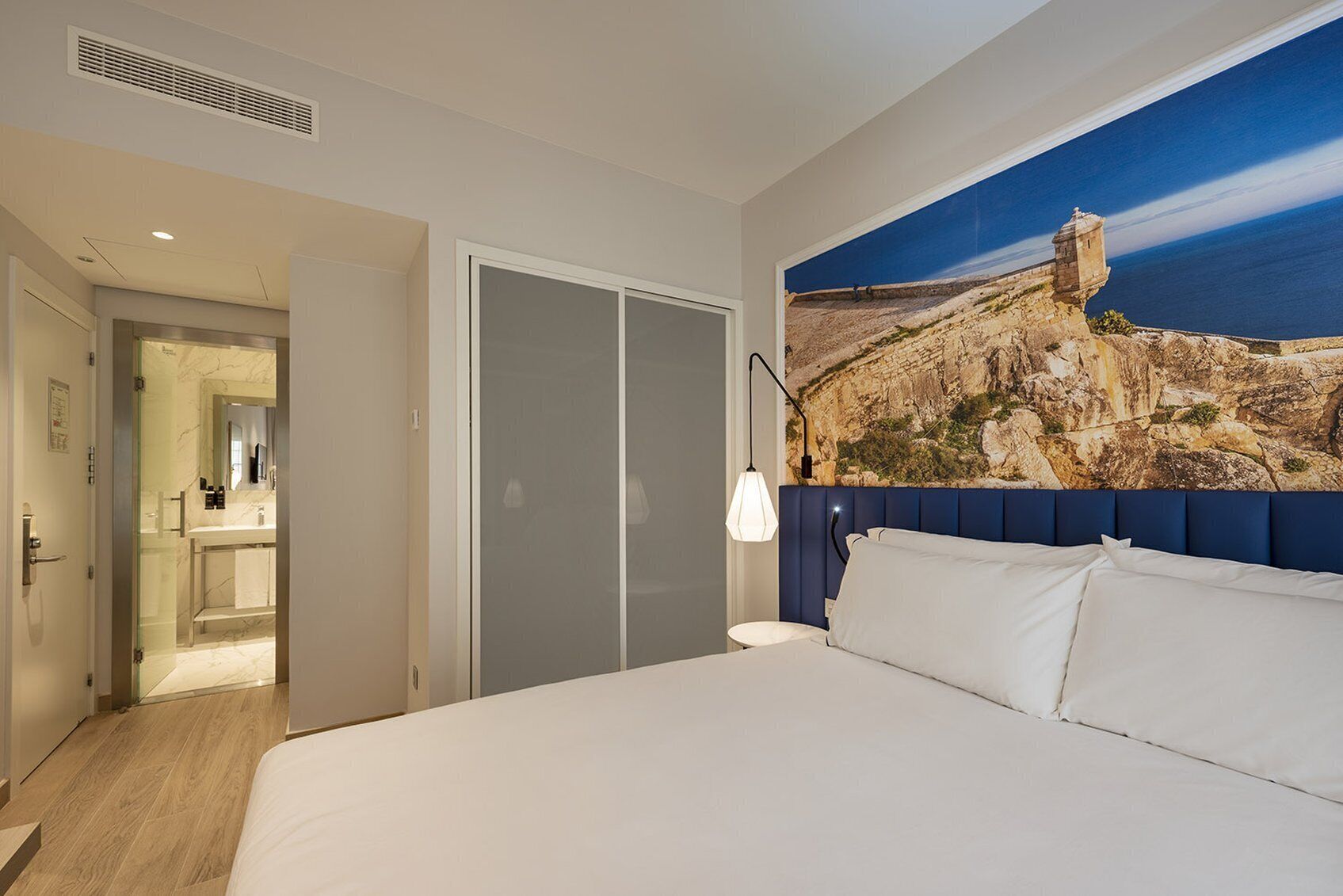 A hotel room with a bed, a bathroom with a glass door, and a large painting of a coastal landscape.