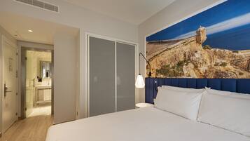 A hotel room with a bed, a bathroom with a glass door, and a large painting of a coastal landscape.