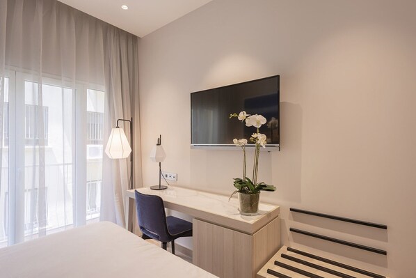 A modern hotel room with a bed, a desk with a chair, a desk lamp, a potted orchid, a flat-screen TV, and a window with curtains.
