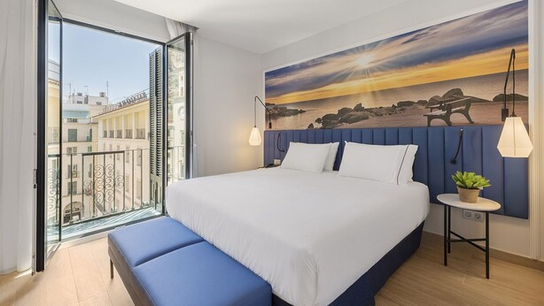 A modern hotel room with a large bed, a bench, a view of the city through a balcony, and a sunset painting on the wall. - Eurostars Mediterranea Plaza (Alicante)