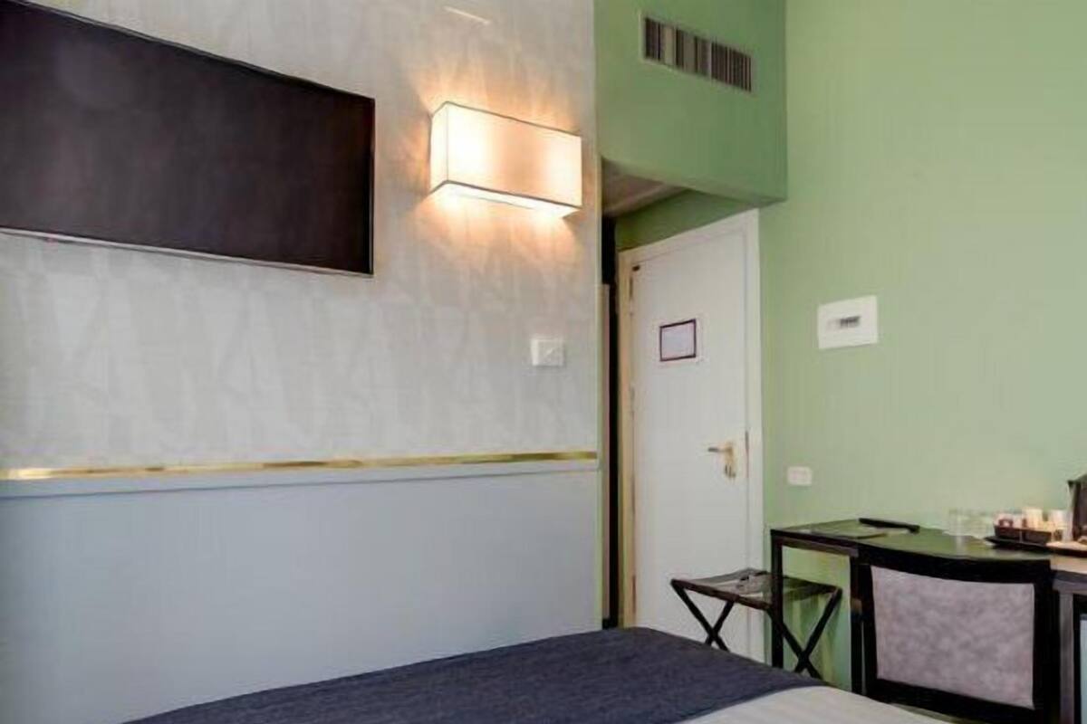 A hotel room with a bed, a desk with a chair, a TV mounted on the wall, and a light fixture above the desk.