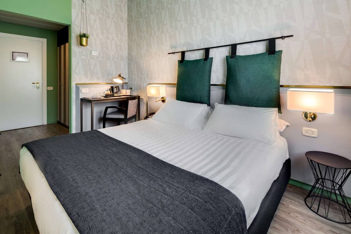 A hotel room with a large bed, a desk with a chair, two bedside lamps, and a small round table.