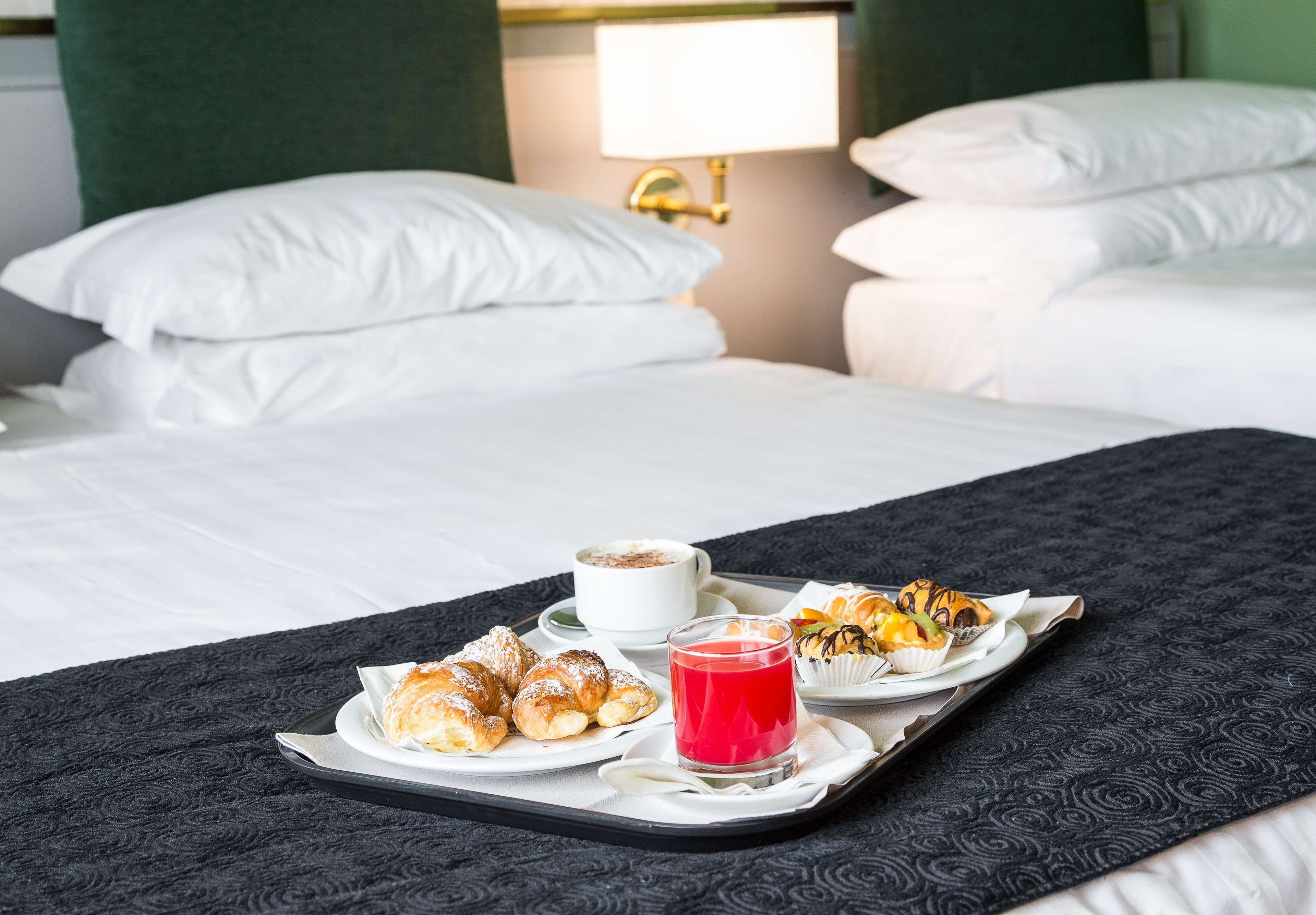 A neatly made bed with white linens, a tray with pastries and a cup of coffee, and a red drink on a black tray.