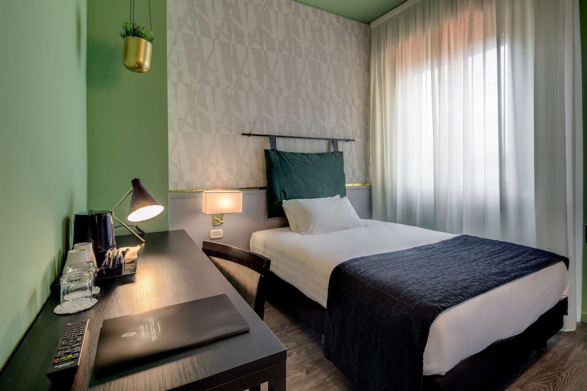 A hotel room with a bed, bedside table, lamp, and a desk with a laptop.