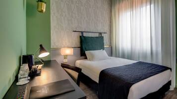 A hotel room with a bed, bedside table, lamp, and a desk with a laptop.