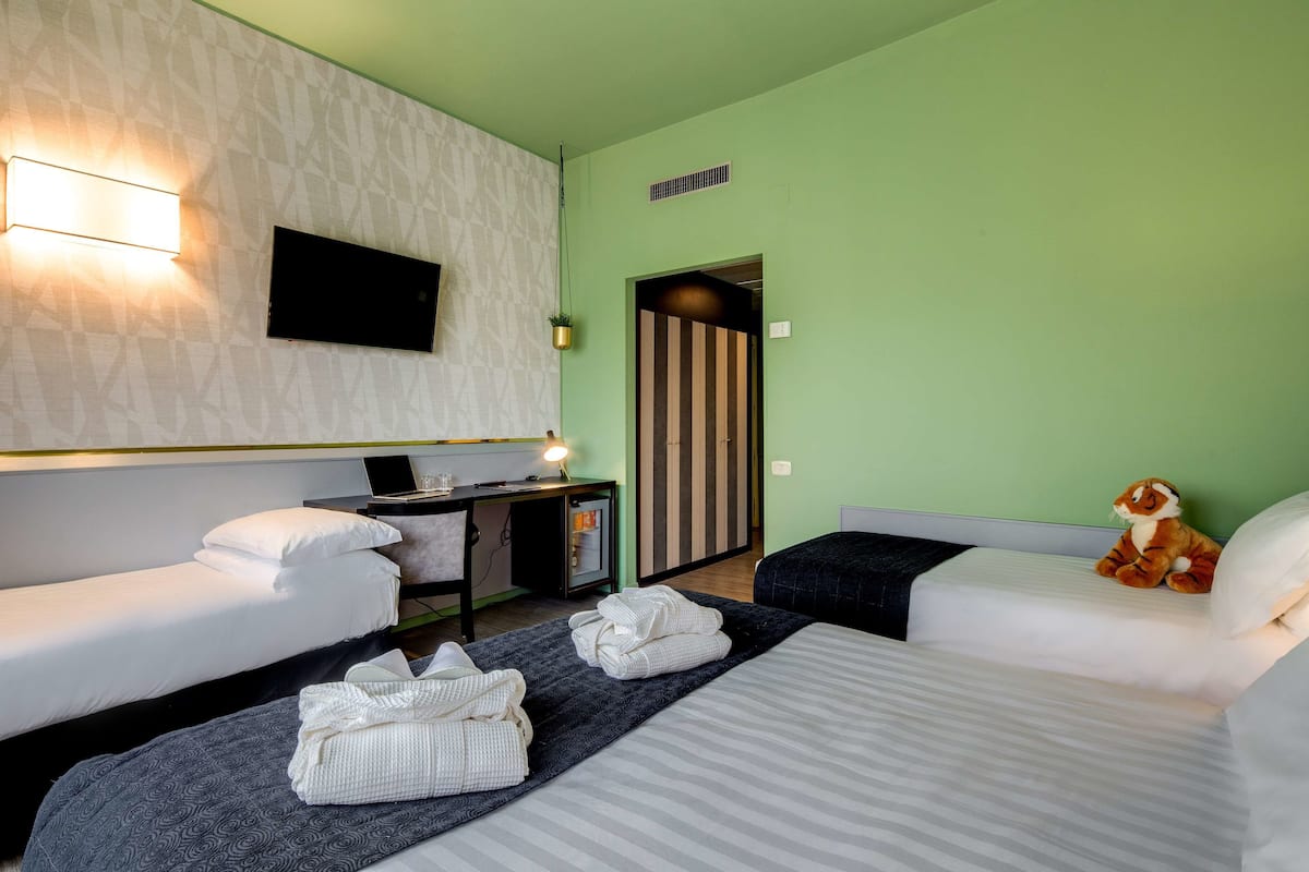 A hotel room with two beds, a desk with a computer, a TV mounted on the wall, and a small plant hanging from the ceiling.