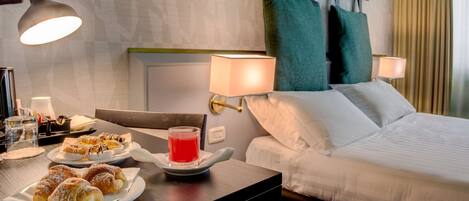 A hotel room with a bed, two bedside lamps, a table with pastries and a glass of red drink, and a view of the city.
