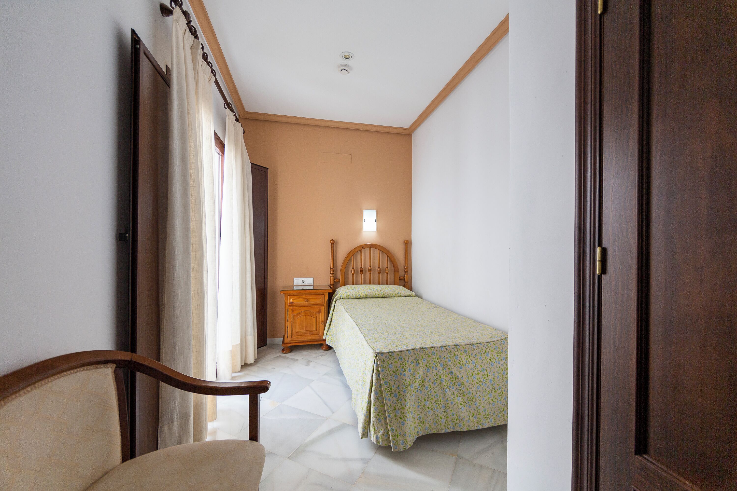 Un dormitorio con cama, mesita de noche de madera, una silla y una puerta.