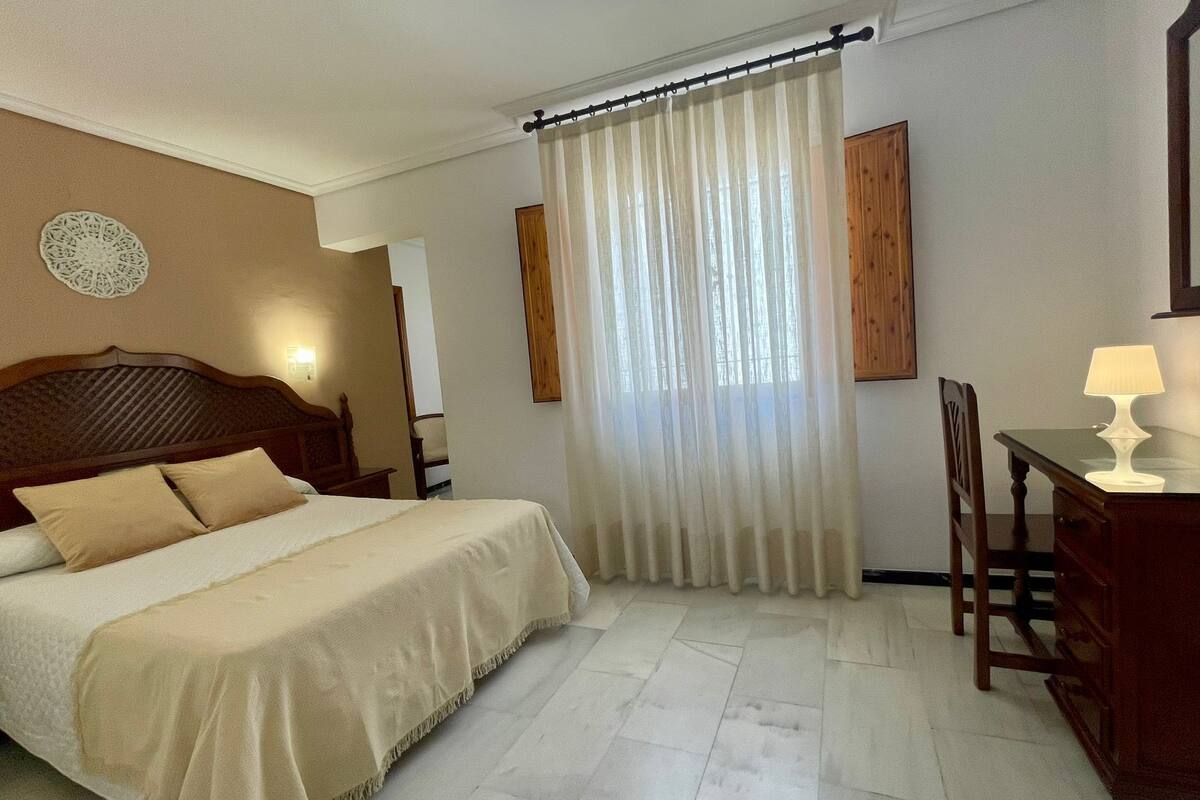 Habitación de hotel con cama, escritorio, silla, lámpara y ventana con cortinas.