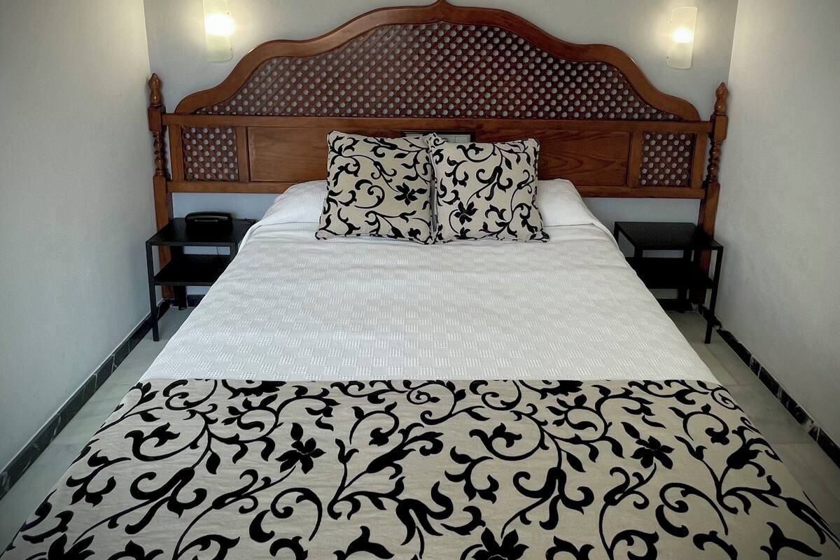 Una cama bien hecha con una colcha estampada, un cabecero de madera con diseño circular y dos lámparas de noche.