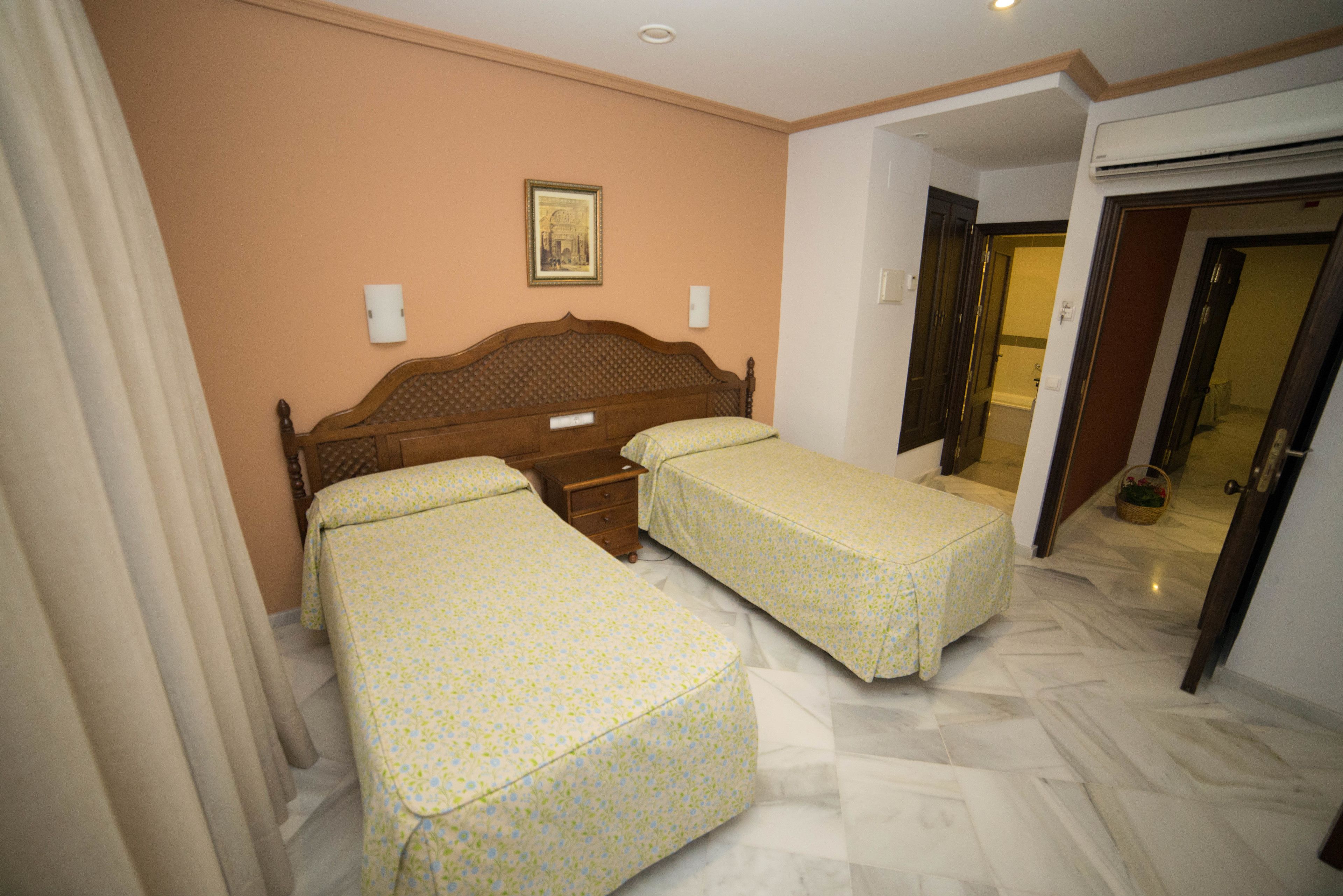 Habitación de hotel con dos camas individuales, un cabecero de madera, una mesita de noche y un cuadro en la pared.