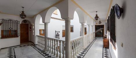 Un corredor amplio con columnas blancas, suelo a cuadros blanco y negro y una escalera al final.