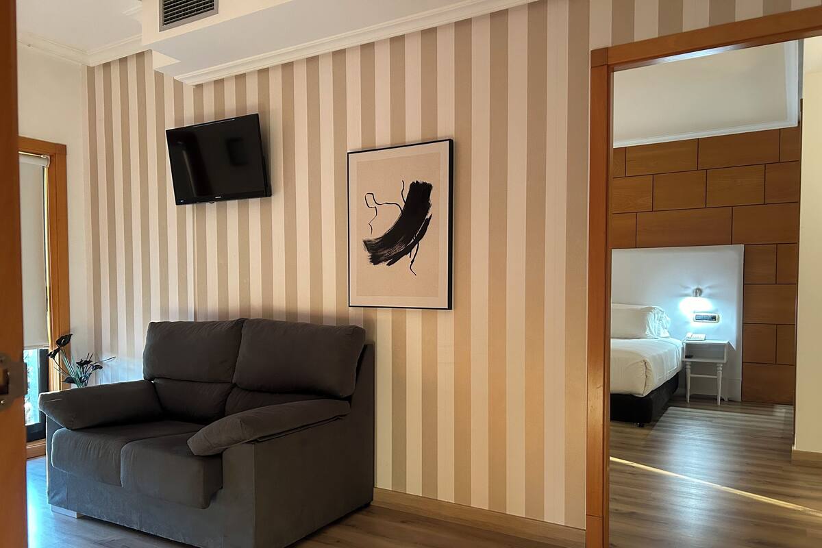 Habitación de hotel moderna con sofá, televisor de pantalla plana, una obra de arte enmarcada y cabecera de madera con mesita de noche y lámpara.