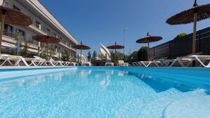 Een zwembad met ligbedden en parasols, een hotelgebouw met balkons, en een helderblauwe lucht.
