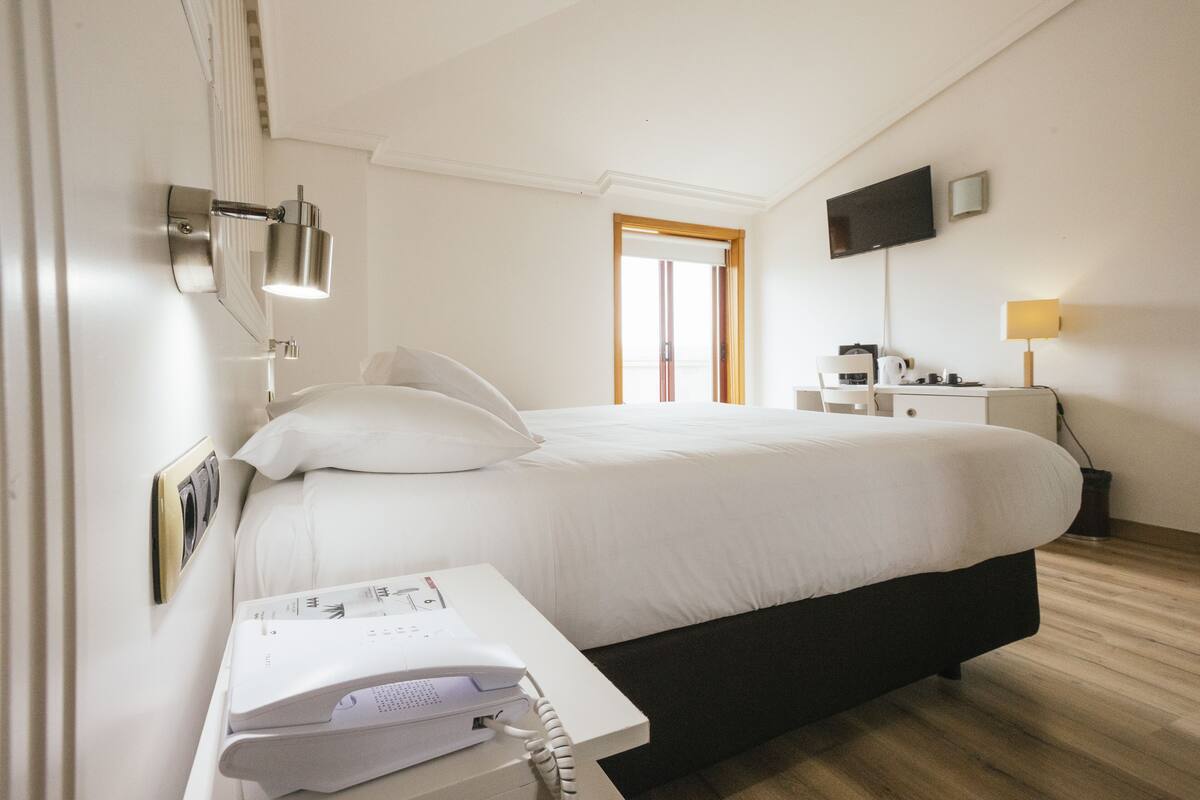 Habitación de hotel con cama, mesita de noche, lámpara, televisión y ventana con vistas.