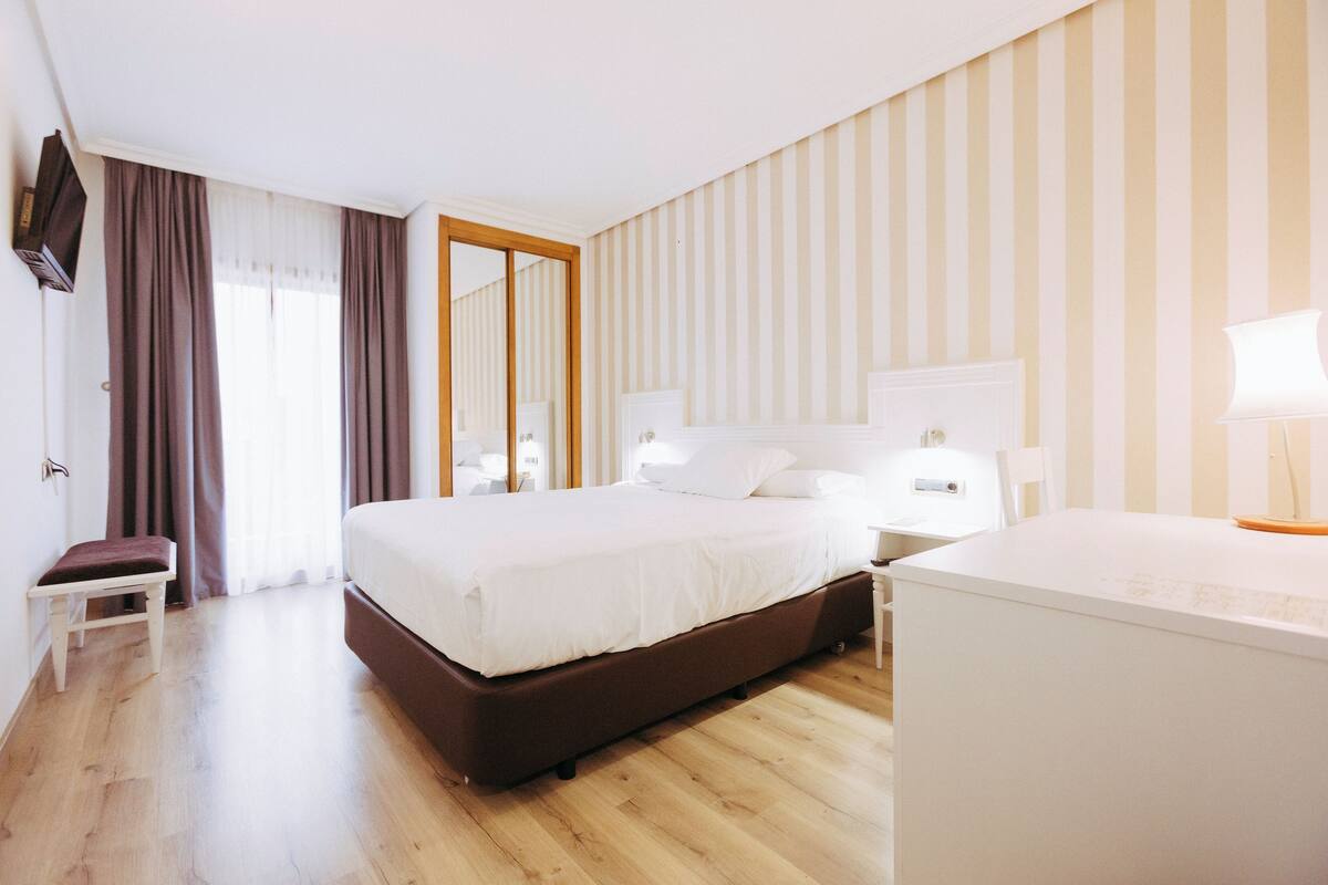 Una habitación de hotel con una cama grande, una mesita de noche, una silla, un escritorio y un televisor.
