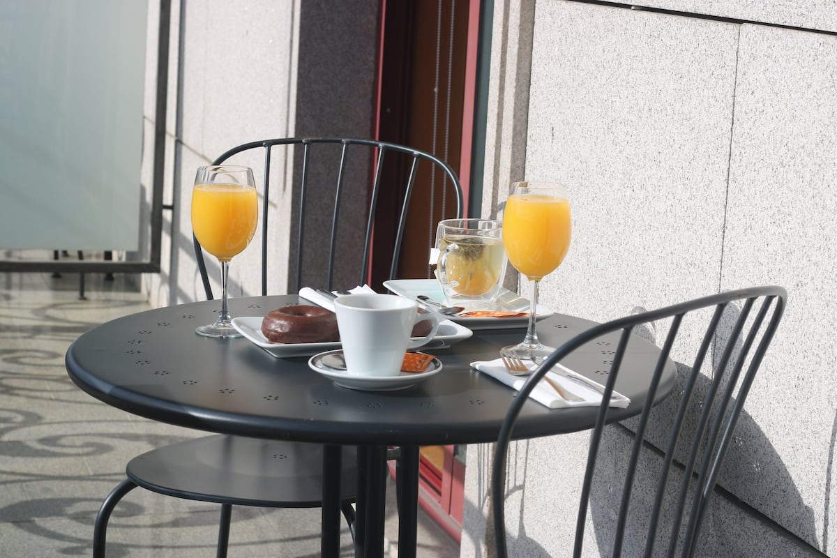 Una mesa metálica redonda con dos sillas, un vaso de jugo de naranja, una taza blanca, un plato con un pastelito y un frasco pequeño de mermelada.