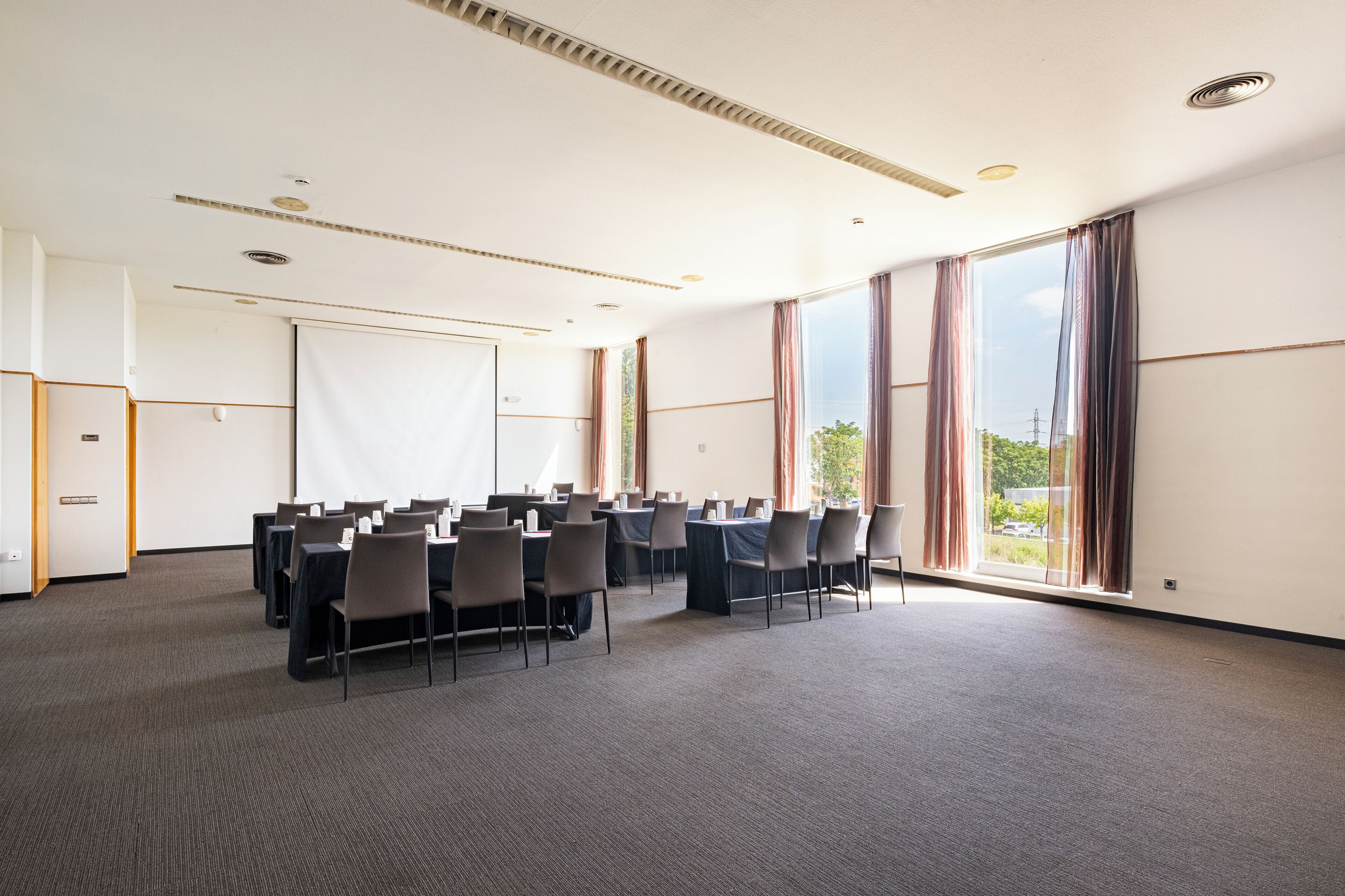 Un salón de conferencias con filas de mesas y sillas, ventanas amplias y una pizarra.