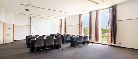 Un salón de conferencias con filas de mesas y sillas, ventanas amplias y una pizarra.