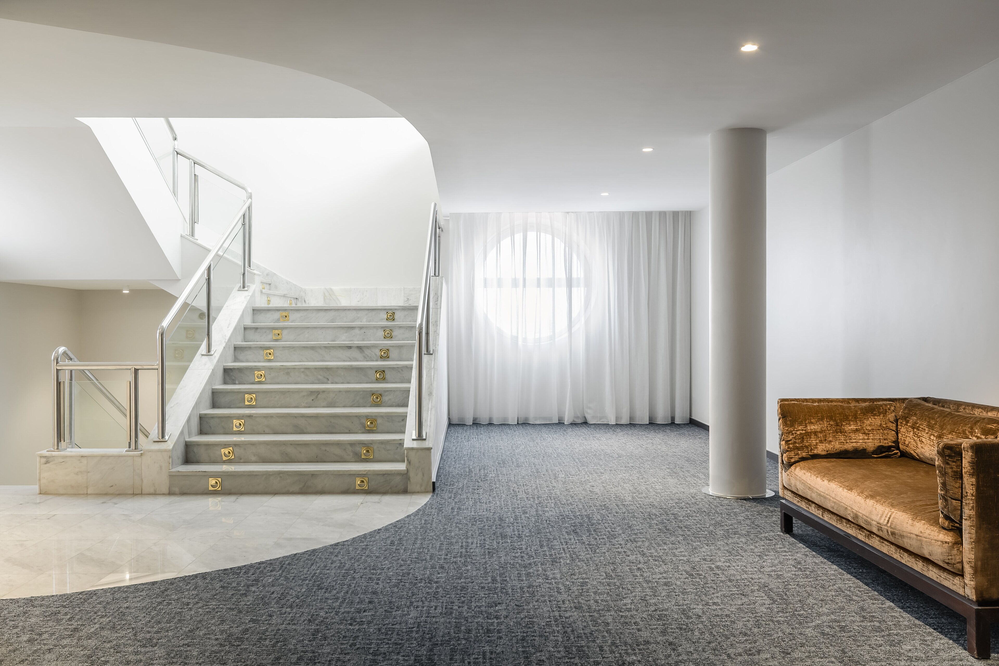 Un interior moderno con una escalera de mármol, piso alfombrado en tono gris, un sofá marrón y un ventanal con cortinas translúcidas.