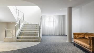 Un interior moderno con una escalera de mármol, piso alfombrado en tono gris, un sofá marrón y un ventanal con cortinas translúcidas.