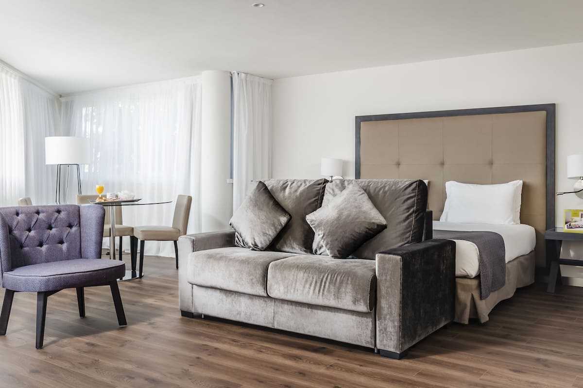 Una habitación de hotel moderna con un sofá gris, un sillón a juego, una cama con ropa blanca y una mesita de noche con lámpara.