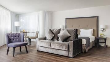 Ein modernes Hotelzimmer mit grauem Sofa, einem passenden Sessel, einem Bett mit weißer Bettwäsche und einem kleinen Tisch mit Lampe.