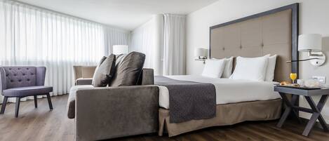 A modern hotel room with a large bed, a sofa, a chair, and a bedside table with a lamp.