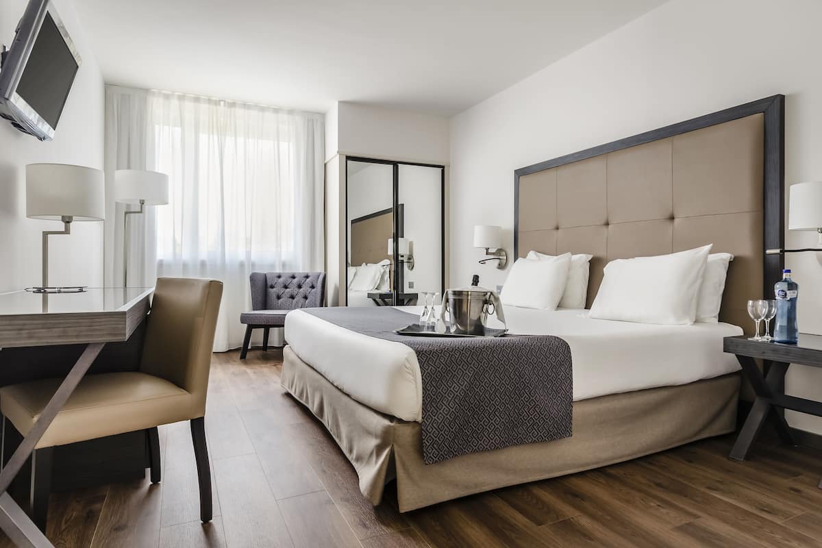 Una habitación de hotel con cama, mesita de noche, lámpara, sillón y escritorio con silla.
