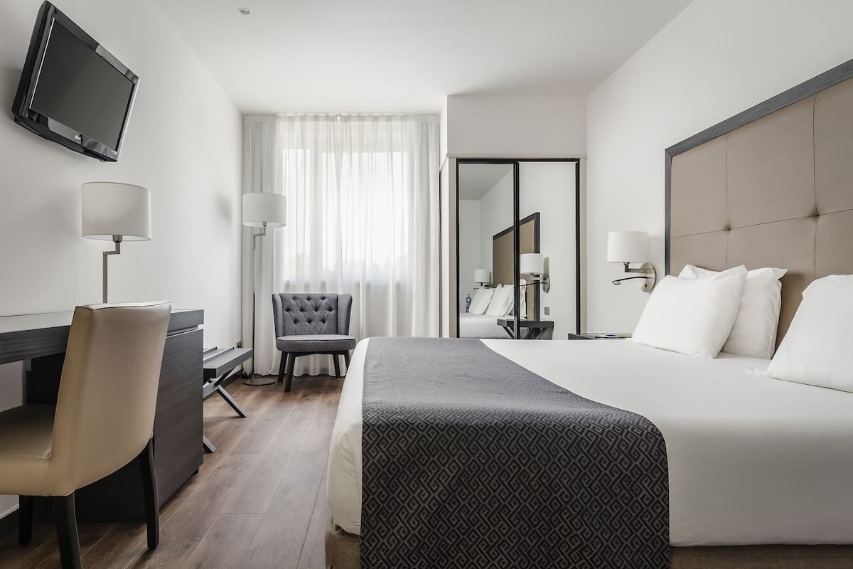 Habitación de hotel con cama, escritorio, silla, televisor y ventana con cortinas.