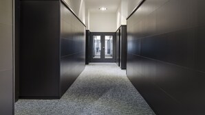 Un couloir moderne aux murs sombres et au sol recouvert d’une moquette, qui mène à une porte en verre au bout.