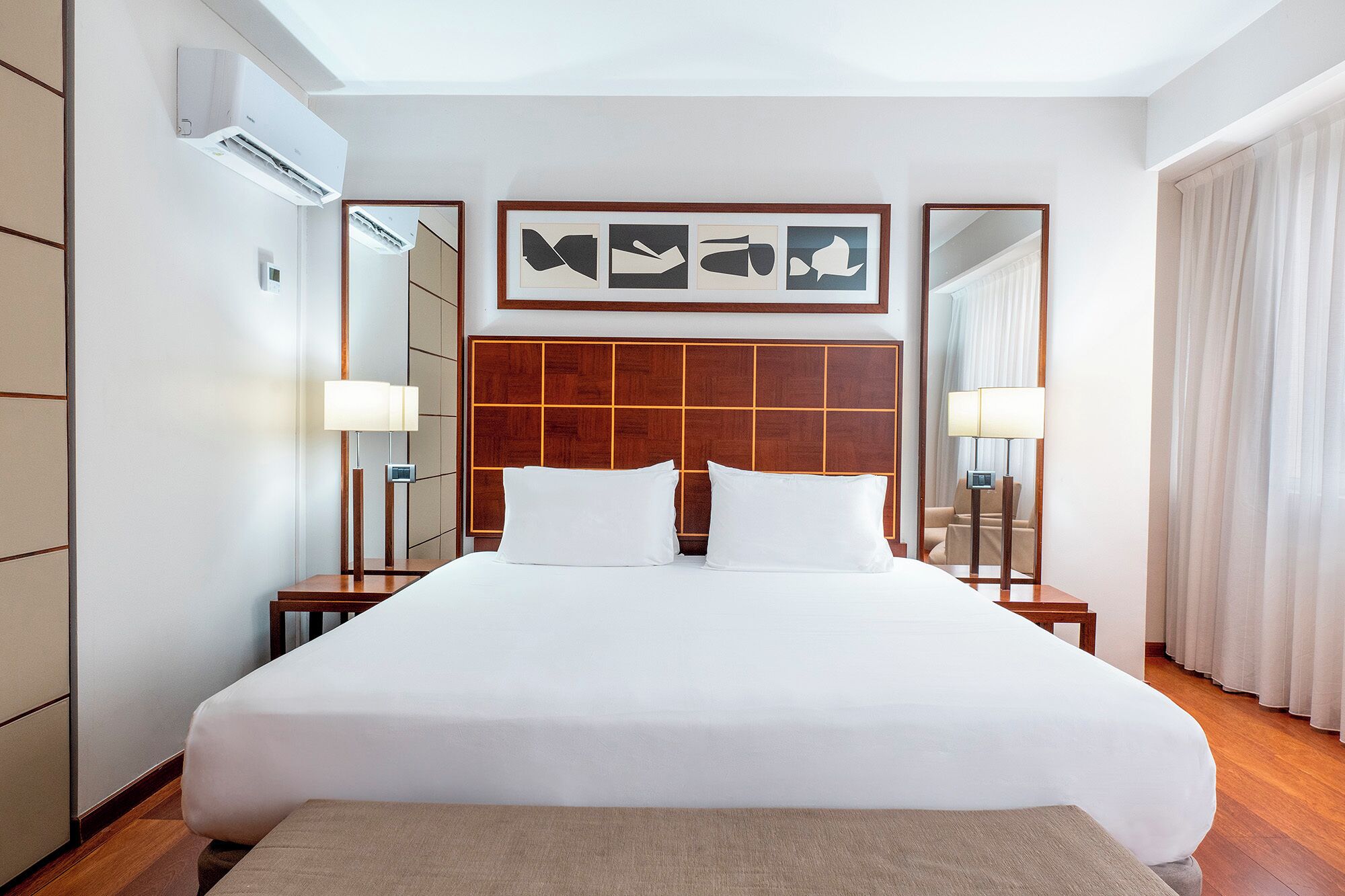 Una habitación de hotel con una cama grande, cabecera de madera, dos mesitas de noche con lámparas, un espejo y una obra de arte enmarcada sobre la cama.
