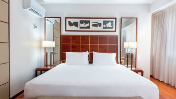 Una habitación de hotel con una cama grande, cabecera de madera, dos mesitas de noche con lámparas, un espejo y una obra de arte enmarcada sobre la cama.
