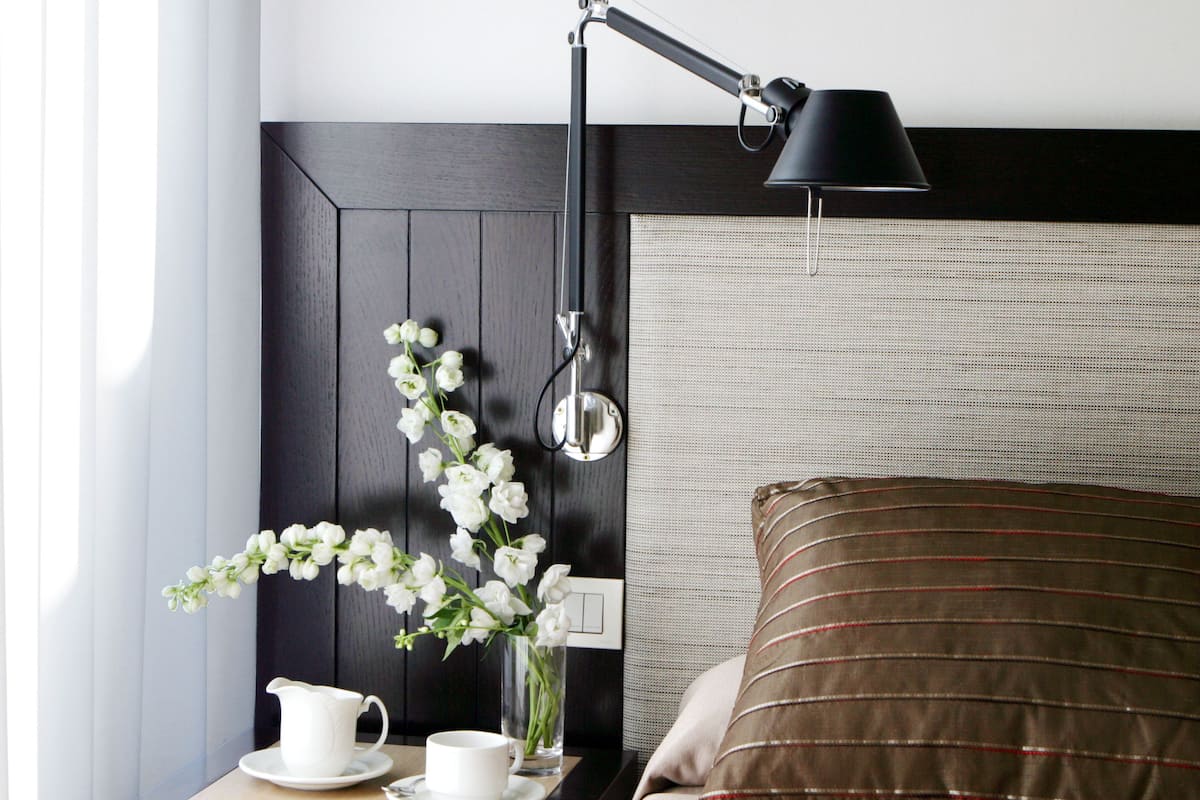 Habitación de hotel impecable con un cabecero de madera oscura, una mesita de noche con lámpara, un florero con flores blancas, una jarra blanca y una almohada a rayas.