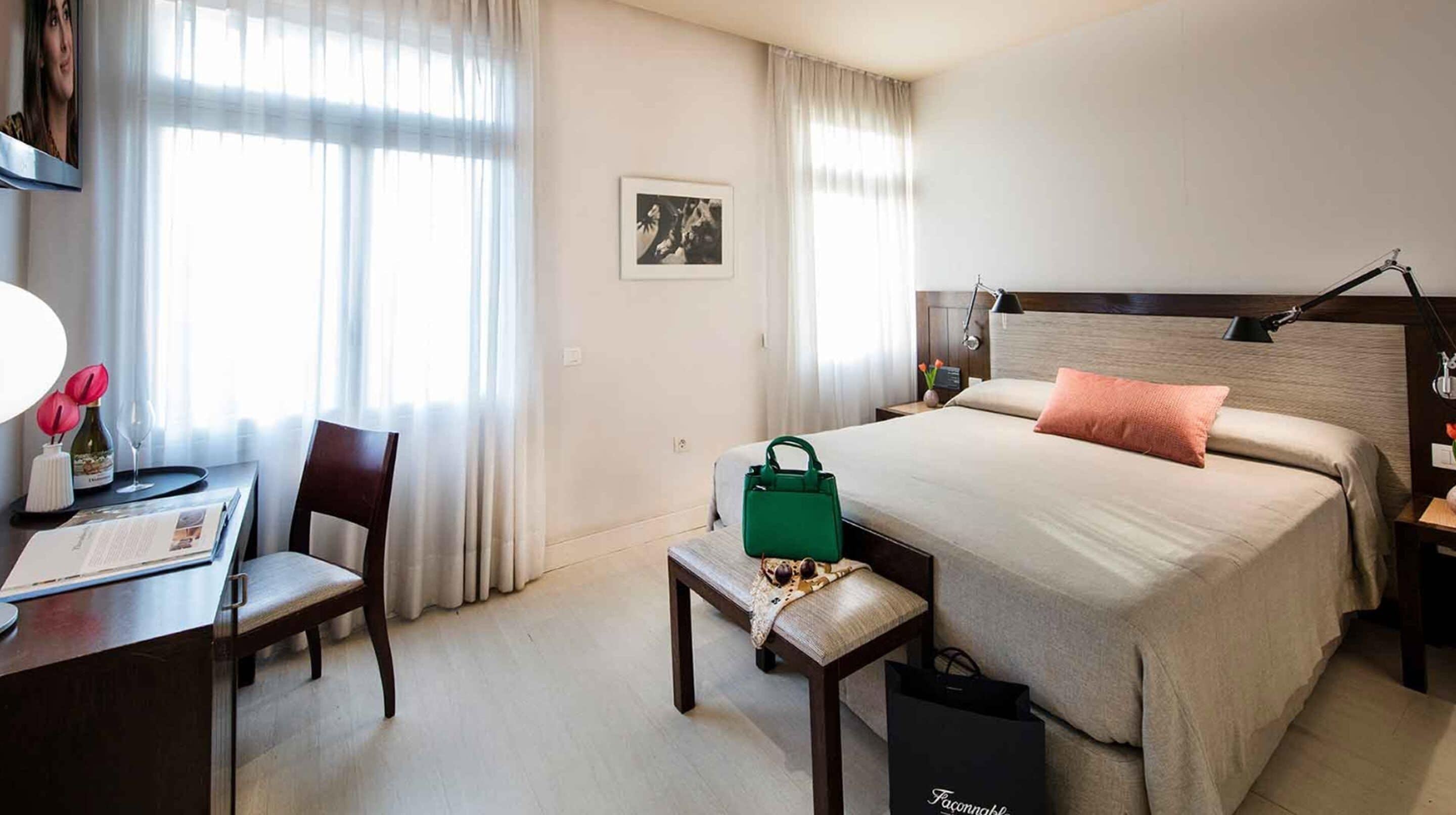 A hotel room with a bed, a desk with a chair, a nightstand, a lamp, and a window with curtains.