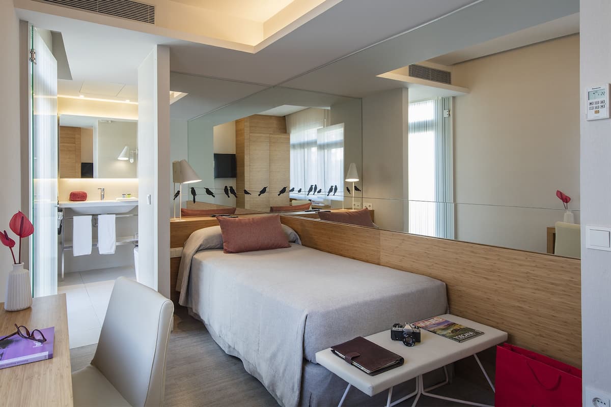Habitación de hotel moderna con cama, mesita de noche y un pequeño escritorio con silla. Hay un espejo en la pared, una ventana con persianas y un baño visible a través de una puerta abierta.