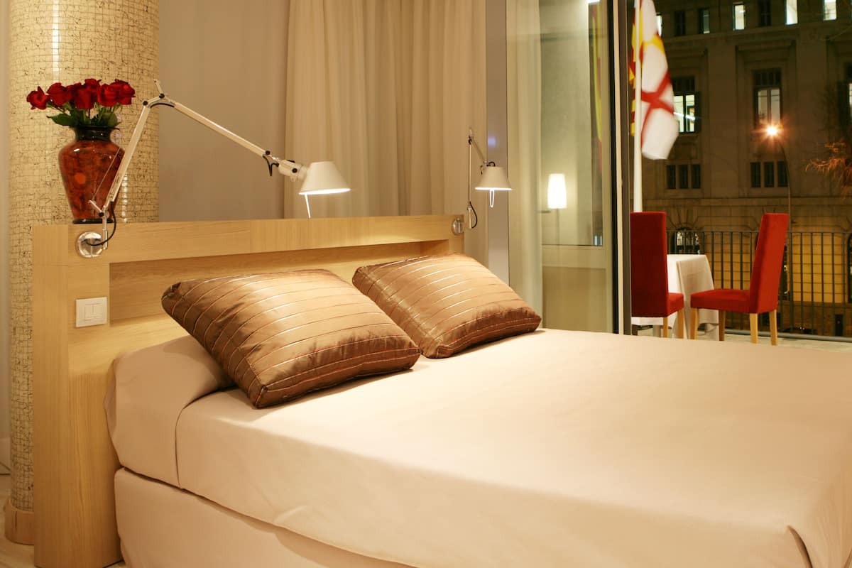 Habitación de hotel con una cama grande, dos lámparas de noche, un jarrón con flores rojas y vistas a la ciudad de noche.