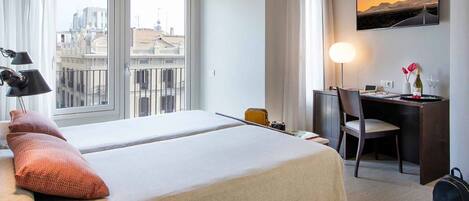 Ein Hotelzimmer mit zwei Betten, einem Schreibtisch, einem Stuhl, einer Lampe und Blick auf die Stadt.