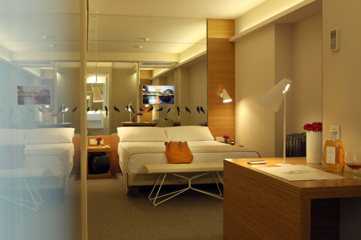 Habitación de hotel con cama, mesitas de noche, escritorio, espejo, televisor y ventana con vista a un paisaje urbano.