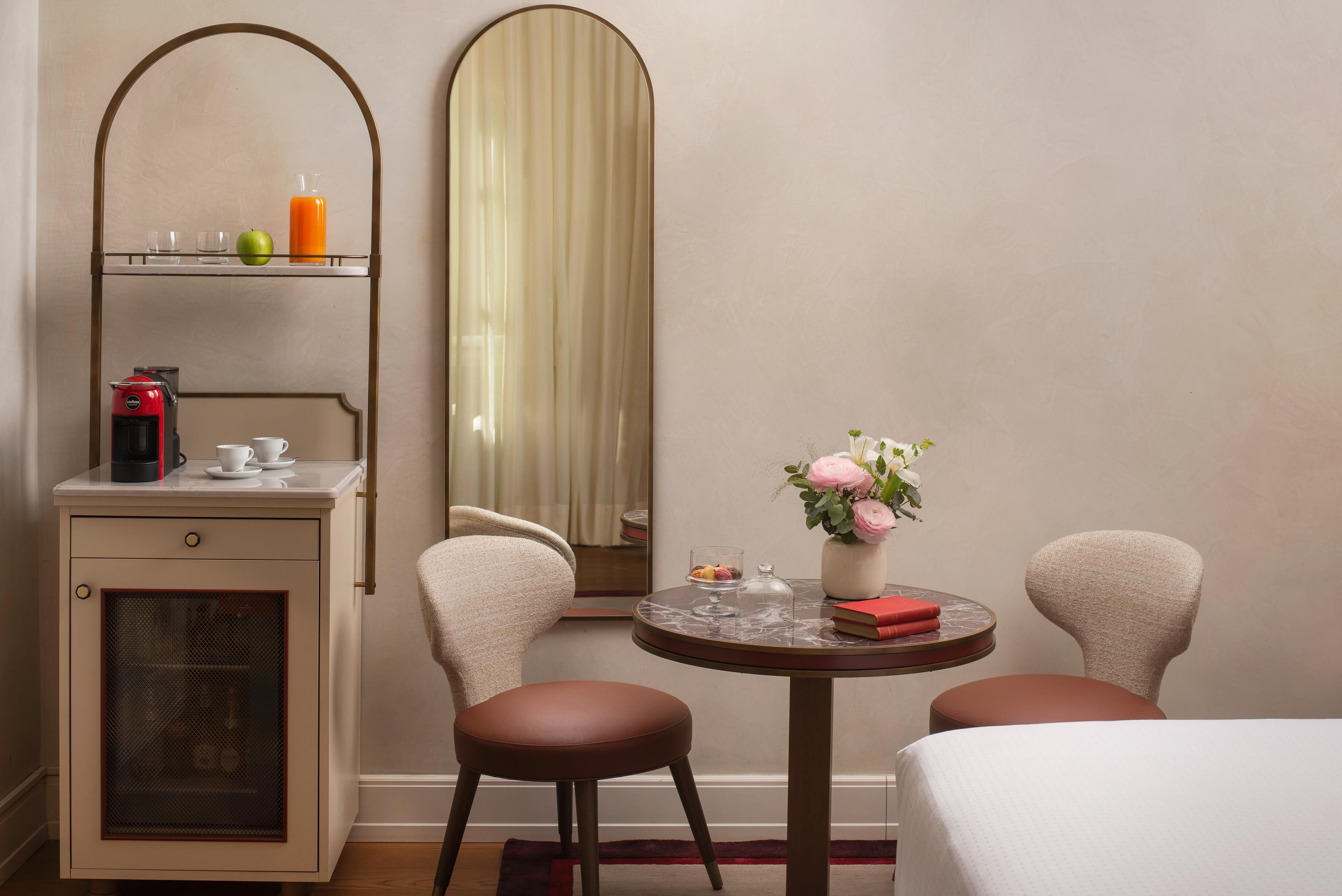 A hotel room with a bed, two chairs, a small table with a vase of flowers, a coffee machine, and a mirror.