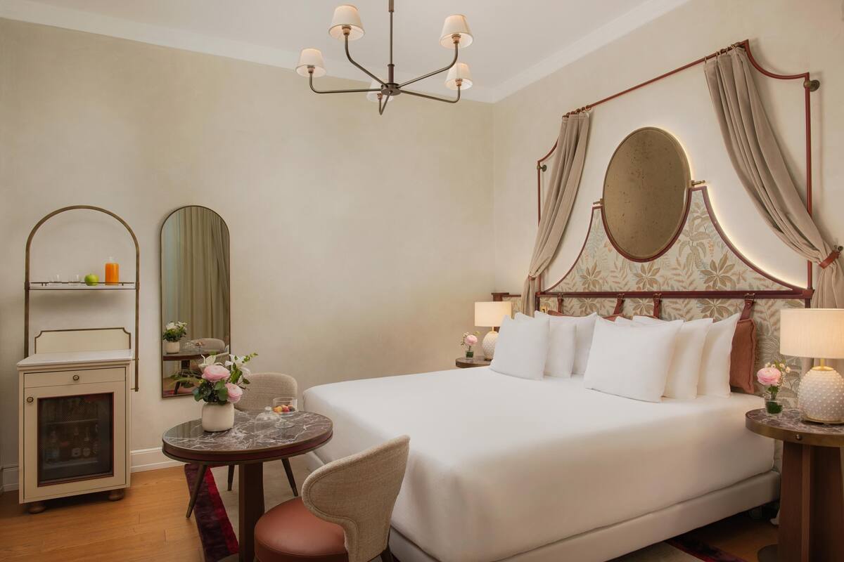 Une chambre d’hôtel avec un grand lit, une table de chevet avec une lampe, une petite table ronde avec une chaise, une armoire et un miroir.