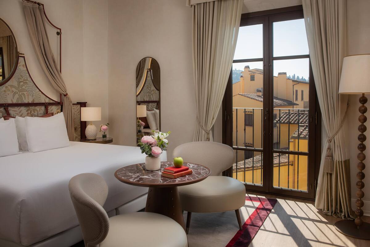 Une chambre d’hôtel avec un lit, deux chaises, une petite table avec un vase et un livre, un miroir et une vue sur le balcon.