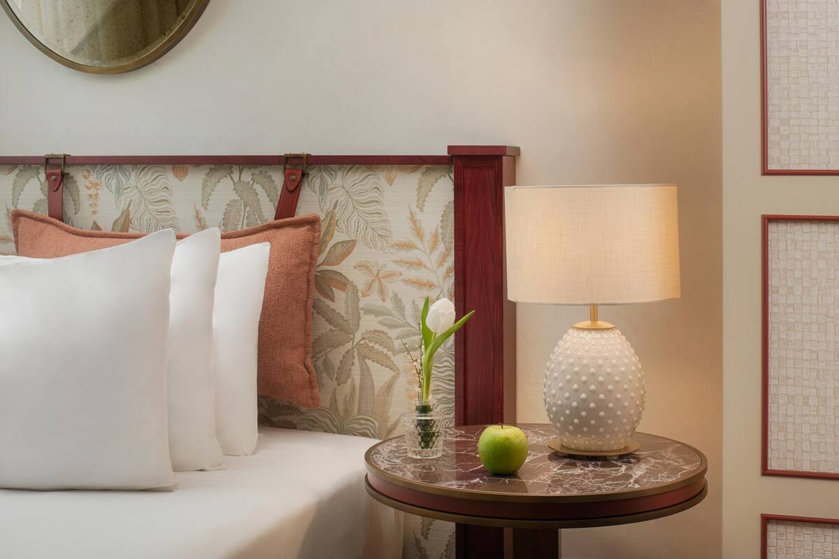 Une chambre d’hôtel bien rangée, avec un lit, des oreillers, une table ronde sur laquelle se trouve une lampe, un vase rempli de fleurs et une pomme verte.