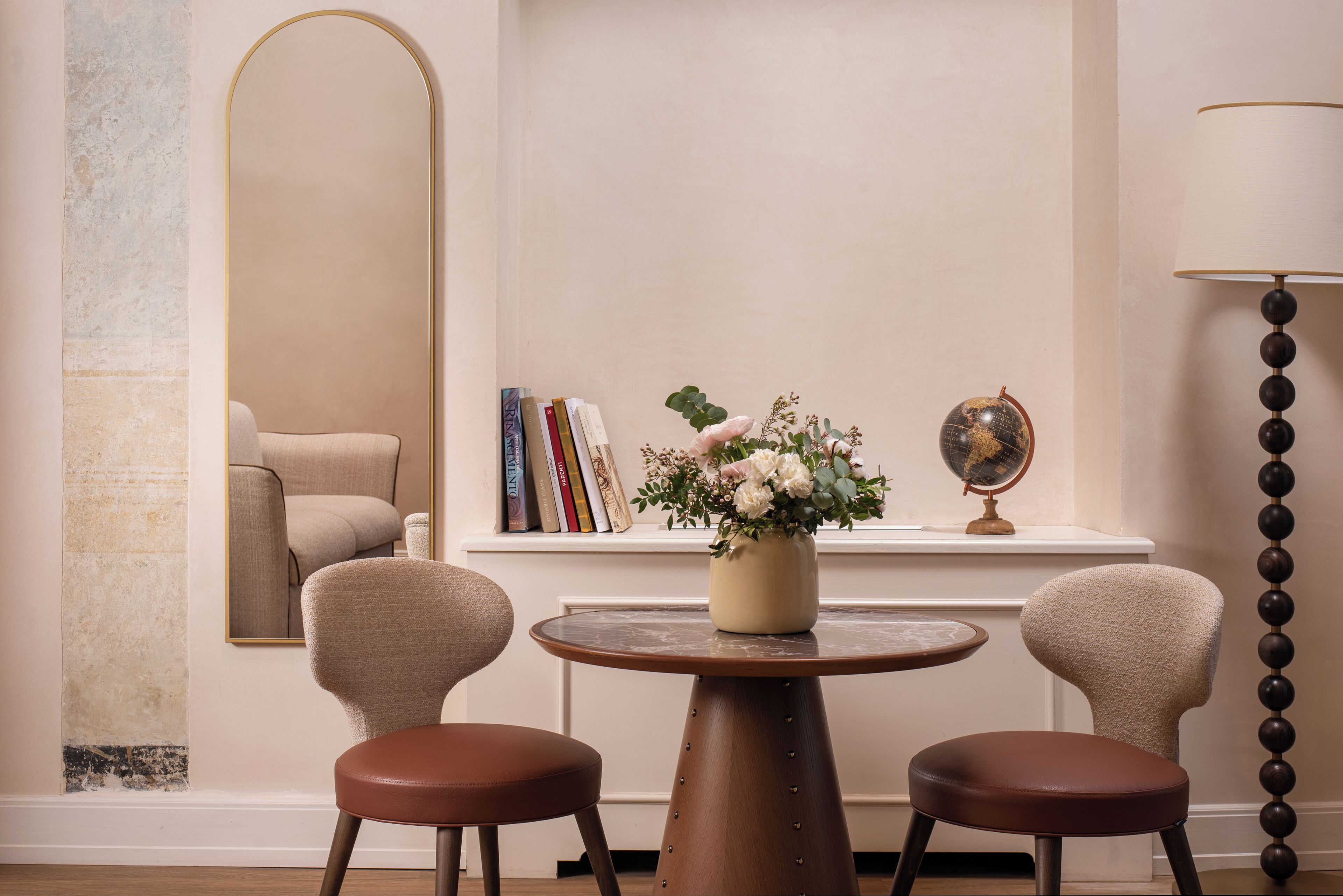 A room with a round wooden table, two chairs, a mirror, books, a globe, and a vase with flowers.