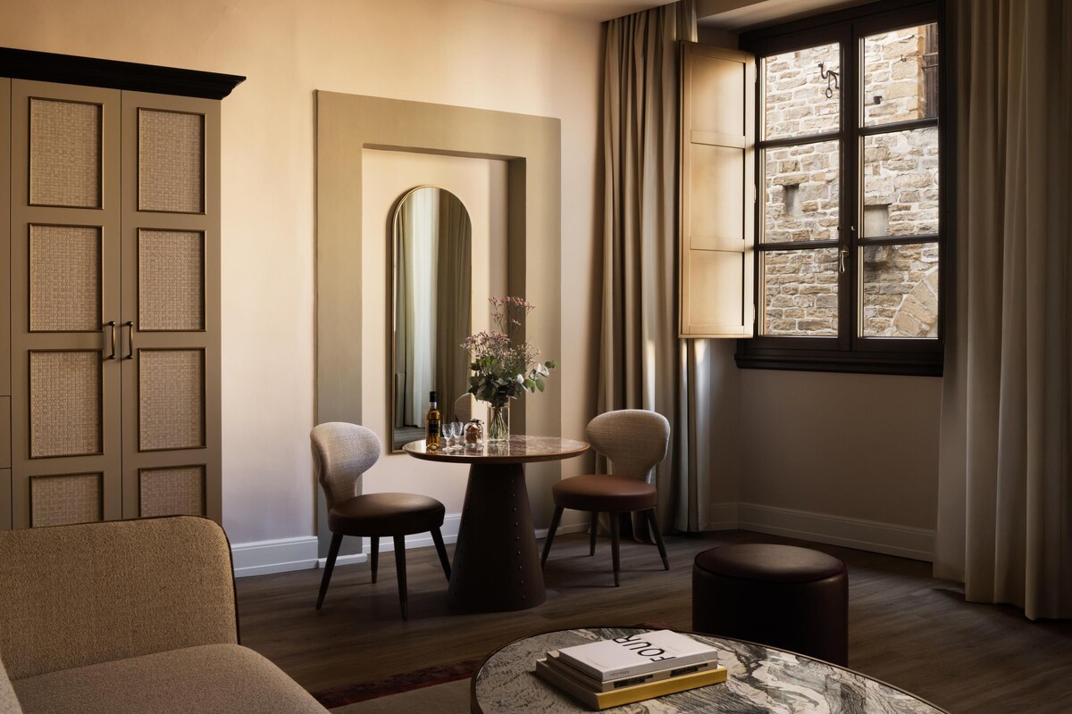Une pièce avec une table ronde, deux chaises, un canapé, un miroir et une fenêtre avec des rideaux.