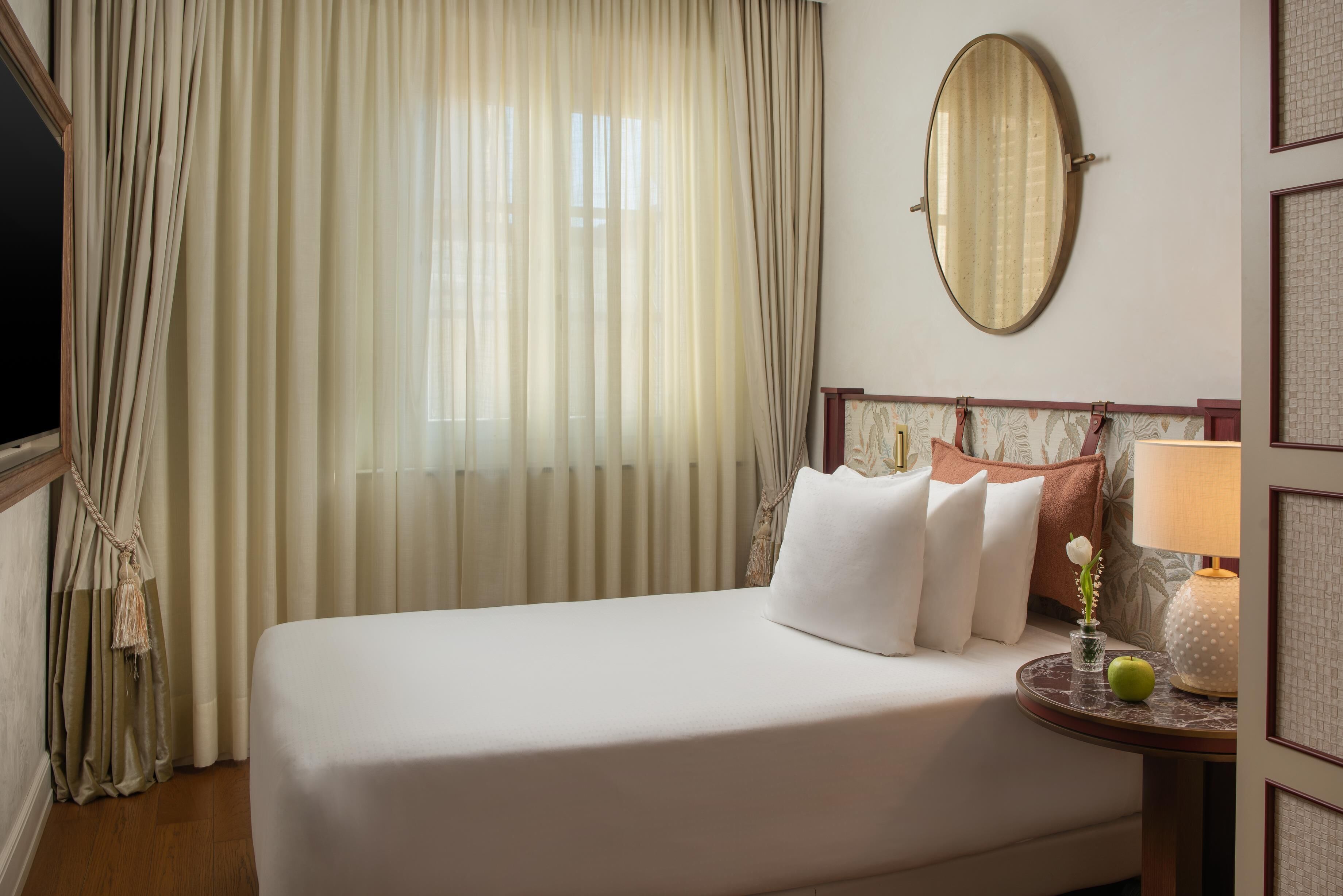 A hotel room with a bed, curtains, a round mirror, a nightstand with a lamp, and a vase with flowers.