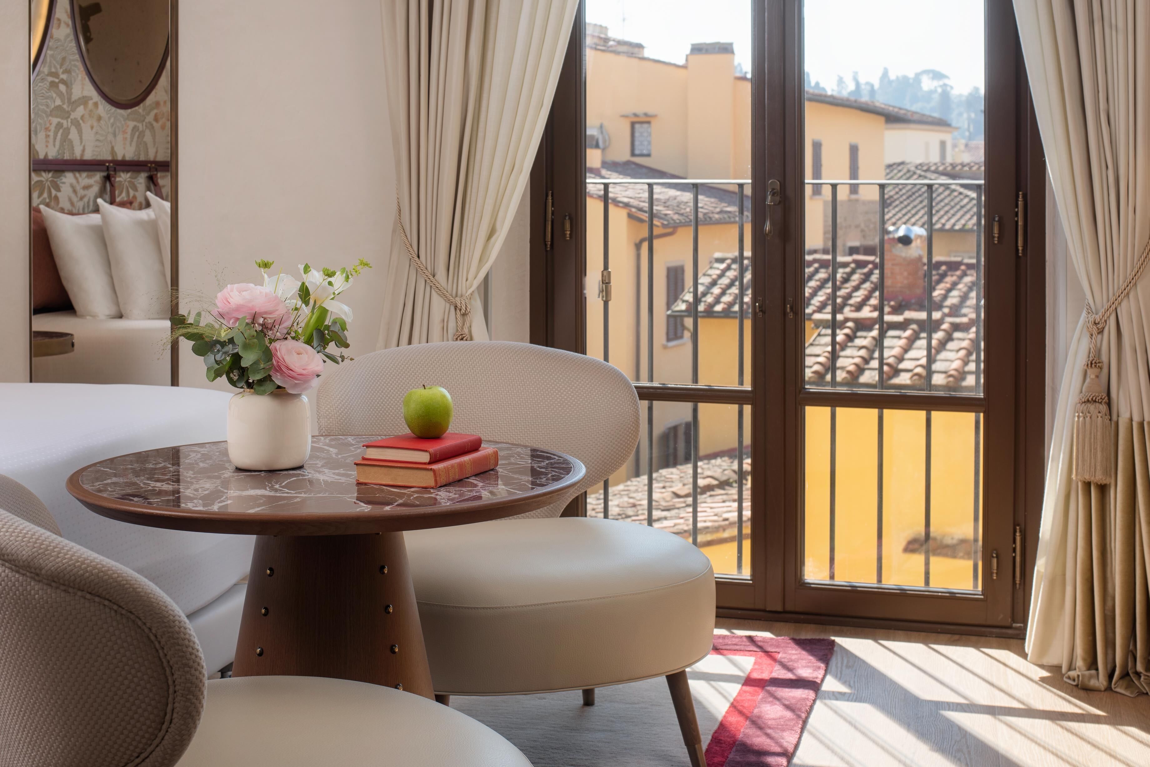 Ein Hotelzimmer mit Balkon, einem runden Tisch mit Blumenvase, einem Buch und einem Apfel.