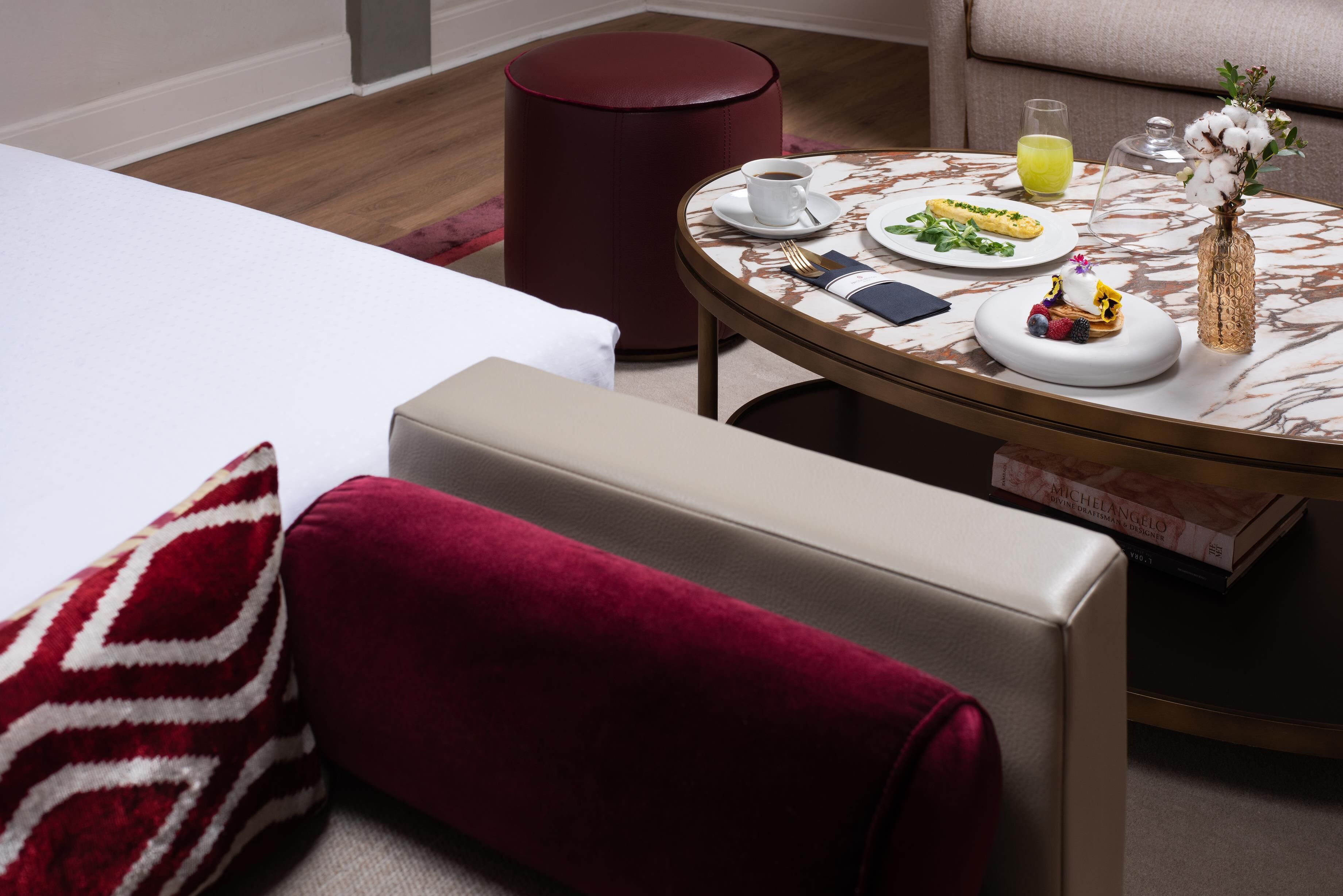 Une chambre d’hôtel avec un lit, une table ronde recouverte d’une nappe blanche, un pouf rouge et un petit vase décoratif.