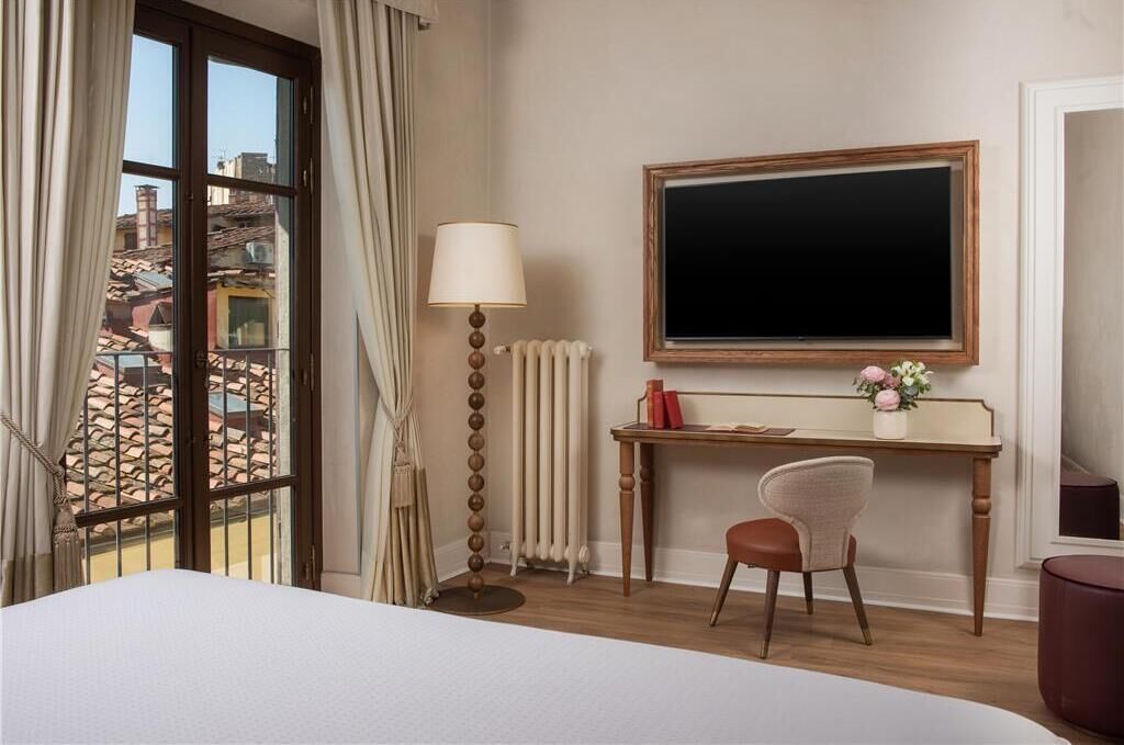 A hotel room with a bed, a desk with a chair, a radiator, a window with curtains, and a view of buildings outside.